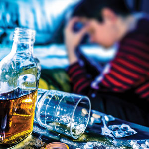 Bottle, tipped over glass, and pills on a table, with a person sitting in the background