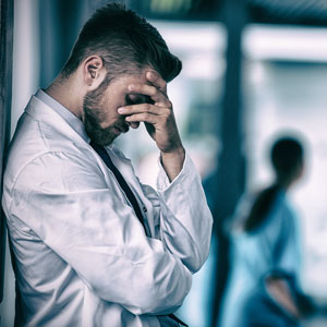 Physician holding his head against a wall with another physician behind him