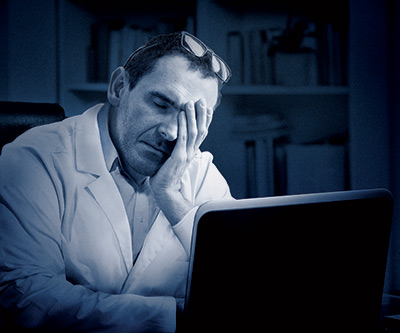 Man sitting at computer holding head in front of bookshelf