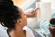 Black woman with hair in tight braids reading book and drinking coffee.