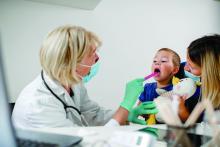 A doctor, wearing a mask, tends to a toddler whose mother is wearing a mask.