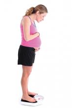 A pregnant woman standing on a scale and peeking over her belly at the number.