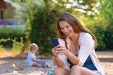 A young mother uses a smartphone while her baby son plays nearby with his toys