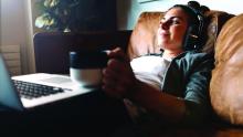 Young woman listening to music from headphones, lying on sofa, using laptop, and drinking coffee.