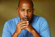 A depressed man sits looking down with his hands in front of his chin.