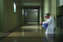 A downcast physician leans against a wall in a corridor.