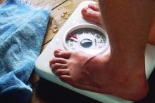 Shown is a close-up of the feet of a man standing on a bathroom scale with a towel lying nearby.