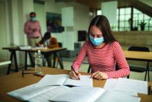 Schoolgirl with protective face mask sitting at desk and writing, at classroom during coronavirus pandemic