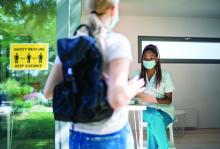 Female teen approaches a check in nurse. Covid safe distancing poster on window.